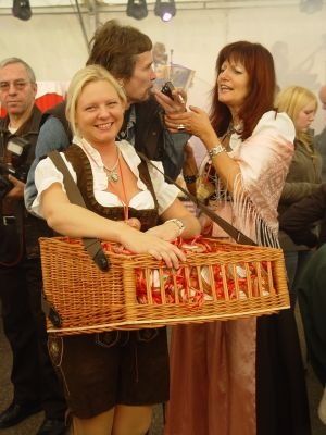 Zwei Frauen in traditioneller deutscher Kleidung, eine hält einen Korb mit roten Süßigkeiten und posiert für ein Foto bei einer Veranstaltung.