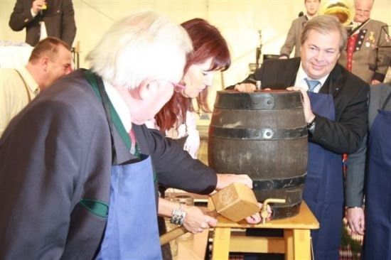 Eine Gruppe von Menschen versammelt sich um einen Fass, wobei einer einen Hammer und ein anderer ein großes Fass hält. Sie scheinen sich auf eine traditionelle Veranstaltung vorzubereiten.