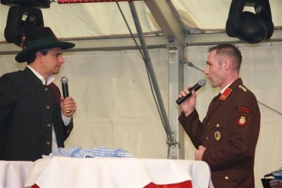 Zwei Männer stehen an einem Podium, einer in einem traditionellen bayerischen Hut und der andere in einer Militäruniform, beide halten Mikrofone und sprechen zu einem Publikum.