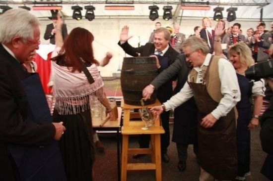 Eine Gruppe von Menschen in traditioneller Kleidung versammelt sich um einen Holzfass, wobei ein Mann ein Getränk in ein Glas gießt. Der Ort scheint ein festliches Ereignis zu sein, mit Scheinwerfern und einer Menge im Hintergrund.