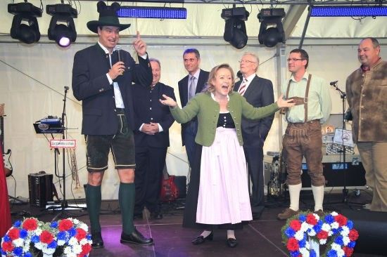 Eine Gruppe von Menschen auf der Bühne, ein Mann in Anzug und Hut mit Mikrofon, eine Frau in grüner Jacke und weißem Rock und andere in formeller Kleidung, mit Musikinstrumenten und Blumen im Hintergrund.