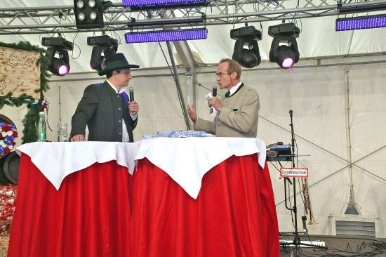 Zwei Männer auf der Bühne, einer hält ein Mikrofon, der andere hält ein Mikrofon und ein Stück Stoff. Sie stehen vor einem Tisch mit roter Tischdecke. Hinter ihnen sind Scheinwerfer und ein Weihnachtsbaum.