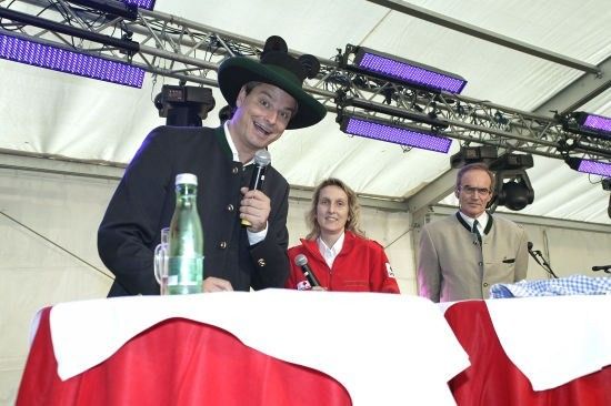 Ein Mann in einem grünen Hut spricht in ein Mikrofon an einem Podium, mit einer Frau in einer roten Jacke und einem weiteren Mann hinter ihm.