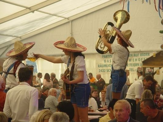 Drei Musiker in Sombreros spielen Instrumente bei einer Outdoor-Veranstaltung, während eine Menschenmenge zuschaut. Einer hält eine Tuba, ein anderer eine Gitarre und der dritte trinkt aus einem Glas.
