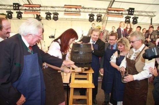 Eine Gruppe von Menschen in traditioneller Kleidung versammelt sich um einen großen Holzfass, wobei einer einen Hammer hält und ein anderer ein Glas.