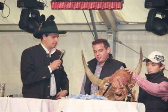 Drei Männer stehen um einen Tisch mit einer großen Kuhkopf-Attrappe. Der Mann auf der linken Seite hält ein Mikrofon und spricht. Der Mann in der Mitte schaut auf den Kuhkopf. Der Mann auf der rechten Seite berührt den Kuhkopf.