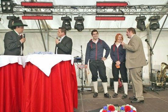 Fünf Personen stehen auf einer Bühne mit einem roten Tischtuch, zwei halten Mikrofone und eine Trompete steht dahinter.