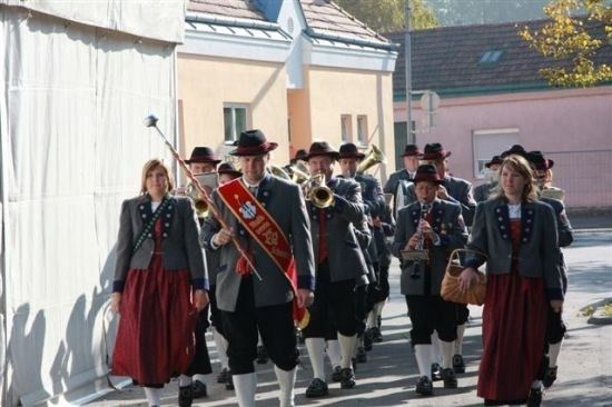 Eine Blaskapelle in traditioneller Kleidung zieht mit Instrumenten spielend und einer Fahne tragend die Straße entlang.