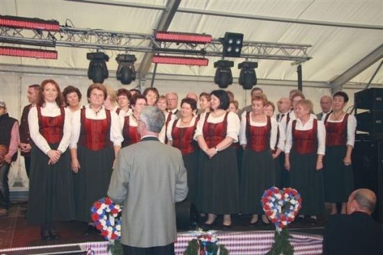 Ein Chor von Frauen in traditioneller Kleidung tritt auf einer Bühne auf, vor ihnen steht ein Mann im Anzug. Die Bühne ist mit Blumen und Kränzen geschmückt.