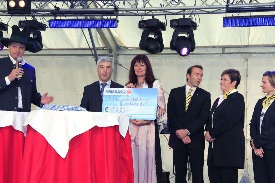 Ein Mann mit Mikrofon steht hinter einem Tisch mit roter Tischdecke, während eine Frau einen großen blauen Scheck mit der Aufschrift 'Sparkasse' und '20 Jahre' hält. Hinter dem Tisch stehen weitere Personen, und an der Decke sind Scheinwerfer montiert.