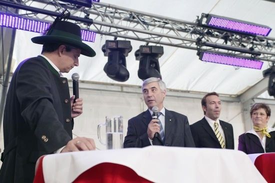 Ein Mann in einem traditionellen Hut spricht in ein Mikrofon an einem Podium, während ein anderer Mann ein Mikrofon und ein Glas Wasser hält. Hinter ihnen stehen zwei Männer in Anzügen und Krawatten, und eine Frau in einem Anzug mit einer gelben Schleife beobachtet.