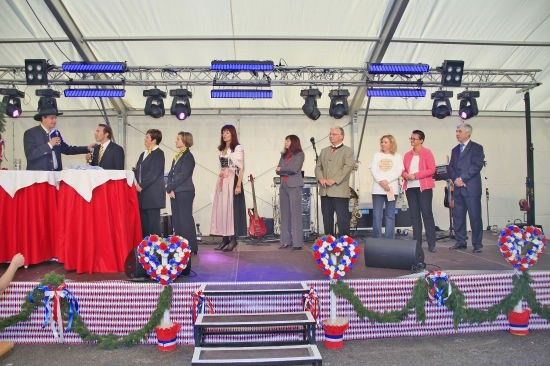 Eine Gruppe von Menschen in formeller Kleidung steht auf einer Bühne unter einem Zelt, mit einem rot-weißen Tischtuch und einem rot-weiß-blauen Kranz.