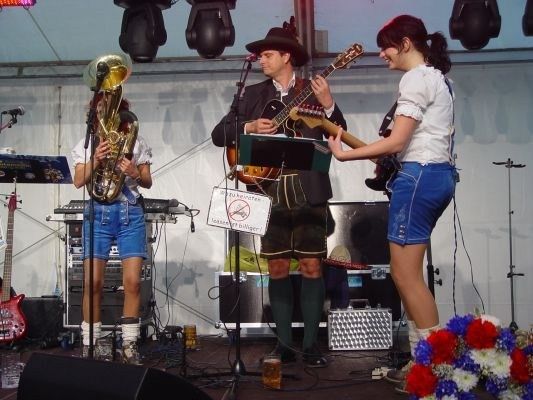 Eine Band spielt auf einer Bühne mit Mikrofon, Gitarren und einem Tuba. Der Mann trägt einen Hut und die Frauen tragen Jeansshorts. Es gibt ein Schild, das 'Kein Rauchen' besagt.