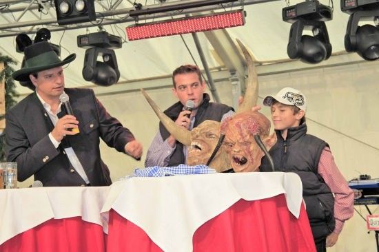 Drei Männer stehen an einem Tisch mit roter Tischdecke. Ein Mann hält ein Mikrofon und spricht, während ein anderer eine Maske mit Hörnern hält. Der dritte Mann, ein Kind, steht neben ihnen.