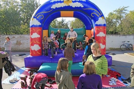 Eine Gruppe von Kindern und Erwachsenen spielt auf einem bunten aufblasbaren Hüpfburg mit Sonne- und Wolkendesign. Einige springen, während andere herumstehen.