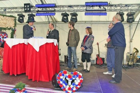 Eine Gruppe von Menschen steht auf einer Bühne mit Mikrofonen, roten Tischdecken und einem Kranz. Im Hintergrund befinden sich Musikinstrumente.