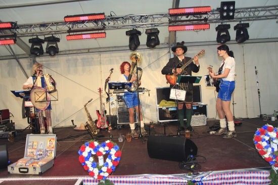Eine Band spielt auf einer Bühne mit verschiedenen Musikinstrumenten, darunter Gitarren, Saxophone und ein Schlagzeug. Die Bühne ist mit rot-weiß-blauen Blumen geschmückt.