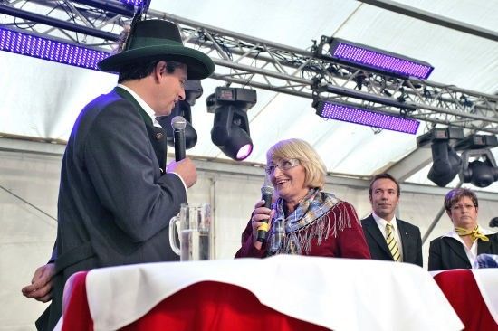 Ein Mann in Anzug und Hut spricht in ein Mikrofon, während eine Frau mit Brille und Schal ein Mikrofon hält und lächelt. Sie sitzen an einem Tisch mit einem Glas Wasser. Hinter ihnen stehen zwei Männer in Anzügen und Krawatten, und über ihnen sind Lichter zu sehen.