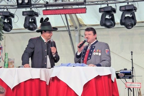 Zwei Männer in traditioneller bayerischer Kleidung sitzen an einem Tisch und singen in Mikrofone. Der Tisch ist mit einer rot-weißen Tischdecke bedeckt, und es gibt Flaschen und ein rotes Schild auf dem Tisch. Scheinwerfer sind an der Decke über ihnen montiert.