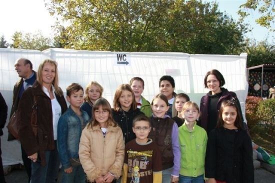 Eine Gruppe von Kindern und Erwachsenen steht zusammen vor einer weißen Wand mit einem WC-Schild. Sie lächeln und scheinen für ein Foto zu posieren.