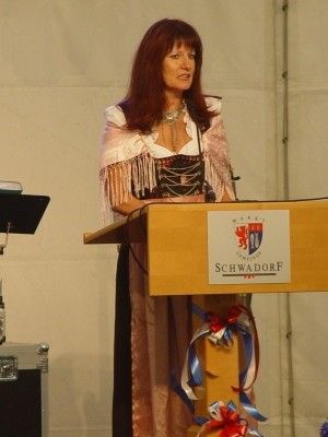 Eine Frau in traditioneller Kleidung steht an einem Rednerpult mit Mikrofon und hält eine Rede. Das Pult hat ein Schild mit der Aufschrift 'Schwadorf' und ist mit rot-weiß-blauen Bändern geschmückt.