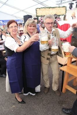 Drei Personen in traditioneller Kleidung, zwei Frauen und ein Mann, posieren mit Bierkrügen bei einer Veranstaltung im Freien.