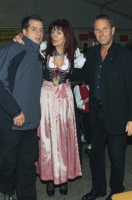 Drei Menschen stehen zusammen, zwei Männer und eine Frau. Die Frau trägt ein traditionelles Kostüm und eine Halskette. Der Mann links trägt eine Jacke und hat seinen Arm um die Frau gelegt. Der Mann rechts trägt einen schwarzen Anzug und schaut in die Kamera.