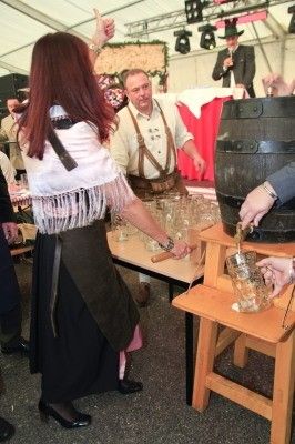 Eine Frau in traditioneller Kleidung schenkt Bier aus einem großen Fass in ein Glas, während ein Mann in traditioneller Kleidung neben ihr steht. Der Hintergrund zeigt andere Menschen, die auf eine festliche Veranstaltung hindeuten.