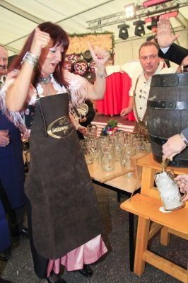 Eine Frau in traditioneller bayerischer Kleidung zapft einen großen Holzfass mit einem Krug an, umgeben von Gläsern und Menschen auf einem Fest.