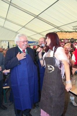 Ein älterer Mann in einer blauen Schürze spricht in ein Mikrofon, während eine Frau in einer grauen Schürze neben ihm steht, beide lächeln und von einer Menge auf einem Outdoor-Event umgeben.