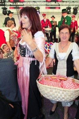 Zwei Frauen in traditioneller deutscher Kleidung tragen einen Korb mit Essen auf einem Festival. Eine Band spielt im Hintergrund.