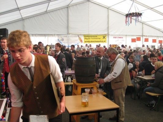 Eine große Menschenmenge versammelt sich in einem Zelt, einige sitzen und andere stehen. Ein Mann in einer Weste schenkt Bier aus einem Fass in ein Glas. Vor ihm steht ein großer Tisch mit Gläsern und einer Speisekarte.