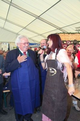 Ein älterer Mann in einer blauen Schürze spricht in ein Mikrofon, während eine Frau in einer grauen Schürze neben ihm steht, beide lächelnd und von einer festlichen Menge umgeben.