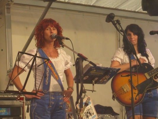 Zwei Frauen treten auf der Bühne auf; eine singt in ein Mikrofon, während die andere Gitarre spielt. Sie sind in übereinstimmenden weißen Blusen und Jeansshorts gekleidet.