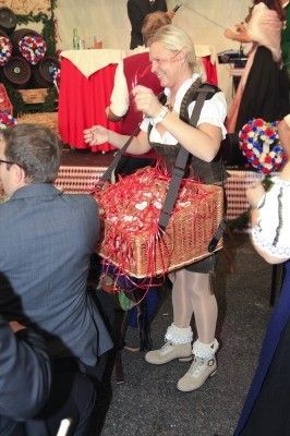 Eine Frau in traditioneller Kleidung trägt einen Korb voller roter Bänder und steht vor einem sitzenden Mann in Anzug. Im Hintergrund ist eine festliche Szene mit Tischen und Fässern zu sehen.