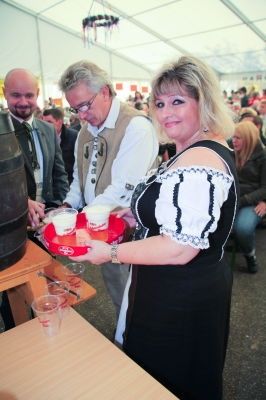 Eine Frau in traditioneller Kleidung serviert Getränke von einem Tablett bei einer Veranstaltung, mit zwei Männern und einer Menge im Hintergrund.