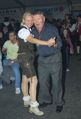Ein älterer Mann und eine Frau in traditioneller bayerischer Kleidung tanzen auf einem Freigelände, während eine Menge zuschaut.
