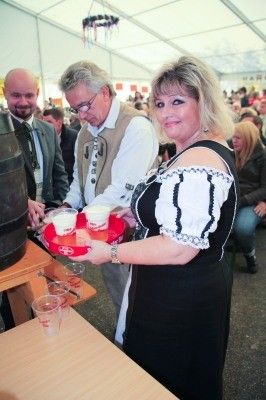 Eine Frau in traditioneller bayerischer Kleidung serviert Getränke von einem roten Tablett bei einer Veranstaltung im Freien, mit zwei Männern und einer Menschenmenge im Hintergrund.