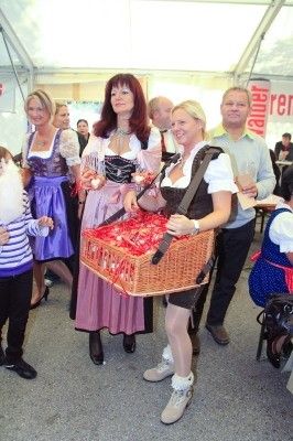Eine Gruppe von Menschen in traditioneller deutscher Kleidung, darunter eine Frau, die einen Korb mit roten Gegenständen trägt, steht in einem großen Zelt. Ein Kind hält ein Plüschtier und ein Mann einen Glas.