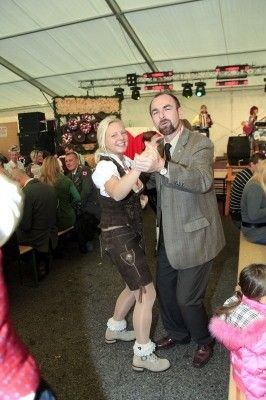 Ein Mann und eine Frau tanzen in einer festlichen Umgebung, während andere Leute sitzen und stehen, und eine Band spielt im Hintergrund.