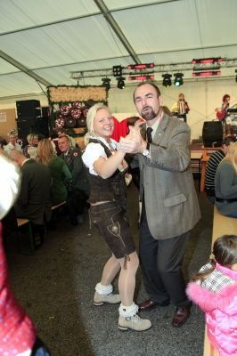 Ein Mann und eine Frau tanzen in einer festlichen Umgebung, möglicherweise bei einer traditionellen deutschen Veranstaltung, mit einer Band im Hintergrund und einer Menge von Menschen, die sitzen und stehen.