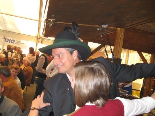 Ein Mann in einem traditionellen bayerischen Hut tanzt mit einer Frau auf einem Festival. Im Hintergrund sind andere Leute zu sehen.