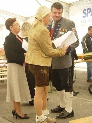 Eine Frau in traditioneller Kleidung hält ein Papier in der Hand, während sie bei einer Veranstaltung im Freien mit einem Mann in Uniform und einer anderen Frau in formeller Kleidung steht.