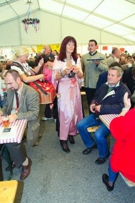 Eine Gruppe von Menschen versammelt sich in einem Zelt, möglicherweise bei einem Festival. Eine Frau in einem rosa Kleid steht in der Mitte und hält ein Glas Bier. Mehrere Personen sitzen an Tischen, und andere stehen und unterhalten sich. Das Zelt ist gut beleuchtet, und es gibt Dekorationen an den Wänden.