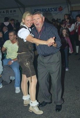 Ein älterer Mann und eine Frau in traditioneller bayerischer Kleidung tanzen zusammen bei einer Veranstaltung im Freien, mit einer Menge im Hintergrund.