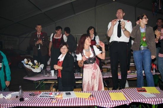 Eine Gruppe von Menschen steht in einem Zelt, einige jubeln und andere stehen um Tische herum. Eine Frau in traditioneller deutscher Kleidung klatscht.