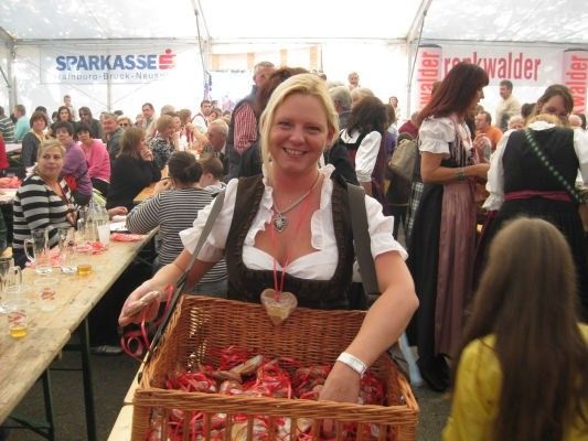 Eine Frau in traditioneller bayerischer Kleidung hält einen Korb mit roten Süßigkeiten und lächelt bei einer überfüllten Veranstaltung. Hinter ihr ist ein Tisch mit Gläsern und einem Banner mit der Aufschrift 'SPARKASSE' zu sehen.