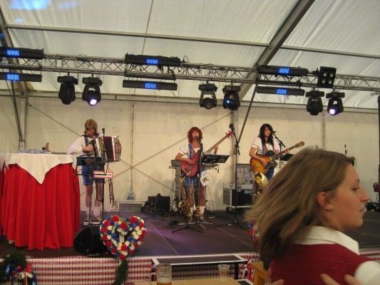 Eine Band spielt auf einer Bühne mit einem roten Tischtuch, Blumen und Scheinwerfern. Die Bandmitglieder spielen Gitarren und Akkordeon. Eine Frau beobachtet sie.