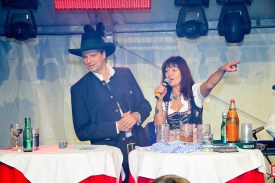Ein Mann und eine Frau stehen an einem Tisch, die Frau singt in ein Mikrofon. Der Mann trägt einen Hut und einen Anzug. Auf dem Tisch befinden sich Flaschen, Gläser und eine Serviette. Der Hintergrund ist unscharf.