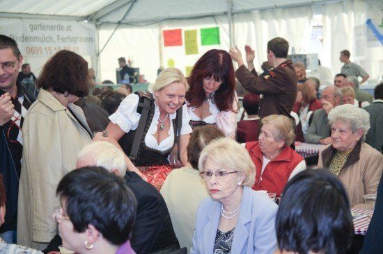 Eine Gruppe von Menschen ist in einem Zelt versammelt, einige stehen und andere sitzen. Eine Frau in traditioneller bayerischer Tracht steht und lächelt. Das Zelt hat Banner und Poster an den Wänden befestigt.